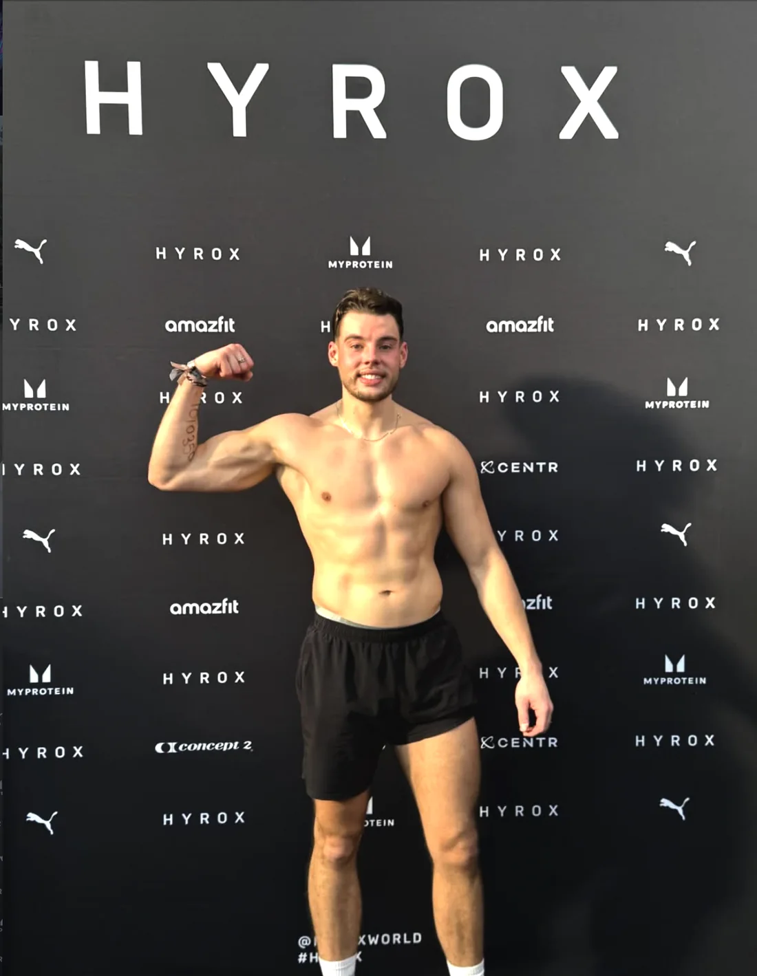 Trinco bij HYROX, lachend, met opgetrokken biceps voor de sponsorwand